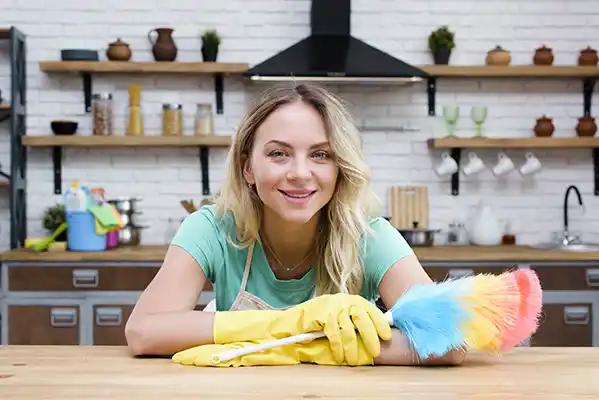 smiling housekeeper leaning kitchen counter holding feather duster looking camera